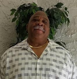 Man smiling in a checkered shirt, standing indoors against a textured wall with a leafy plant behind him