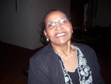 Smiling woman in glasses and black top, seated indoors against a dark background.