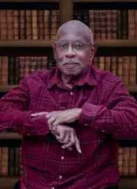Portrait of a man in a red plaid shirt seated before a bookshelf, hands folded, with a calm expression