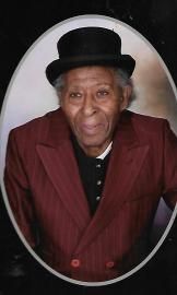Portrait of a person in a black bowler hat and maroon coat, posed against a plain backdrop.