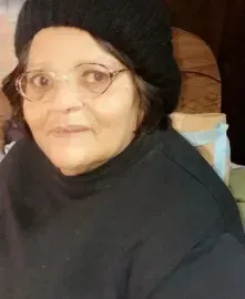 Person wearing glasses and a black headscarf, seated indoors against a wooden background.