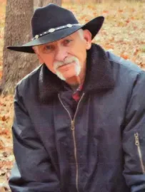 Man in a black cowboy hat and jacket sitting outdoors against a rocky background