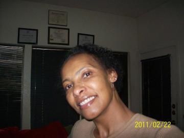 Woman smiling and tilting her head indoors, with framed certificates on the wall behind her.
