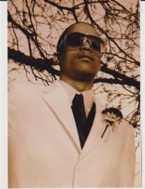 Man in a white suit and dark sunglasses, standing outdoors beneath tree branches.