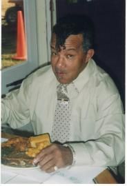 Man in a light shirt and tie seated at a table, eating a meal indoors.