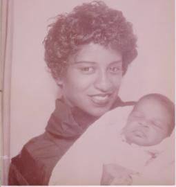 Woman in a dark top holding a sleeping baby wrapped in a white blanket, softly tinted pink.