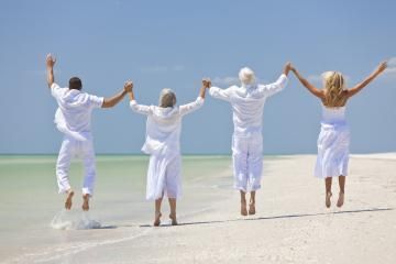 Four people in white clothes jump on a sunny beach, holding hands by the sea.