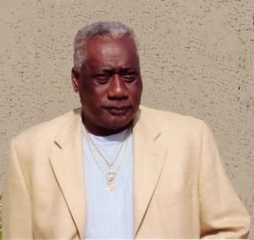 Man in a cream blazer and white shirt posing against a beige wall, looking off to the side.
