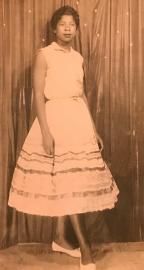 Person posing in a light dress and heels in front of a curtain backdrop.