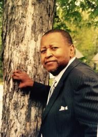 Man in a dark pinstripe suit leaning against a large tree outdoors, looking at the camera.