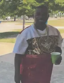 Person outdoors holding a green cup, wearing a white and red shirt with a gold chain.