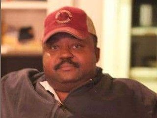 Man in red cap and dark jacket sitting indoors, looking at the camera with a neutral expression.