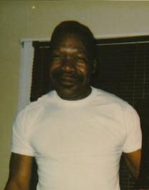 A man in a white T-shirt standing indoors in front of a dark window blind.