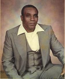 Studio portrait of a man in a light gray suit seated against a soft brown backdrop
