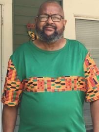 Man standing on a porch, wearing a green shirt with colorful patterned sleeves, smiling at the camera.