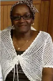 Smiling woman wearing glasses and a white crocheted shawl, seated indoors against a wood-paneled wall.