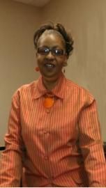 Woman in an orange striped shirt and glasses sits against a beige wall, facing the camera.