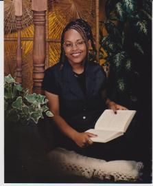 Smiling woman seated with an open book in a tropical-style setting, surrounded by plants and woven decor.