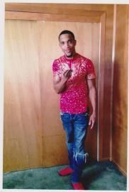 Person standing indoors against a wooden door, wearing a red patterned shirt and jeans, holding up one finger.