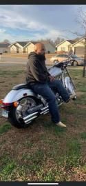 Person sitting on a silver motorcycle in a grassy suburban yard, wearing a black jacket and jeans.