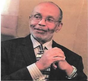 Man in a dark suit and striped tie, smiling with hands clasped against a warm indoor background