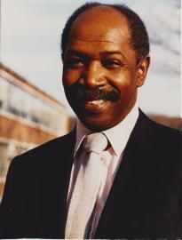 Portrait of a smiling man in a dark suit and tie outdoors, with a blurred building in the background