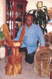 Man sitting indoors beside two carved wooden animal sculptures and a glass display cabinet