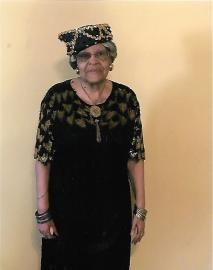 Person standing indoors against a plain wall, wearing a black floral dress and matching headscarf.