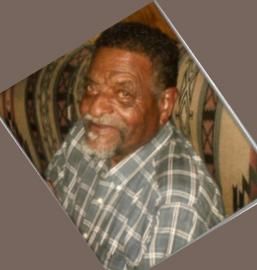 Smiling man in a gray plaid shirt seated on a patterned couch, shown in a rotated photo frame