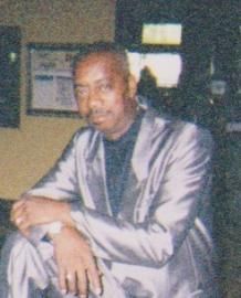 Man in a silver suit sitting indoors, resting his arms on his knee and looking at the camera.