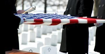 Hands holding a red, white, and blue ribbon in front of rows of white gravestones outdoors