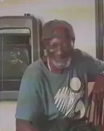 Man smiling in a teal T-shirt and cap, leaning on a chair indoors.