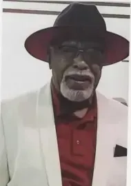 Man in black hat and glasses, wearing a white jacket over a red shirt, standing indoors.