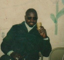 Man in dark suit and sunglasses seated indoors, holding a yellow cup