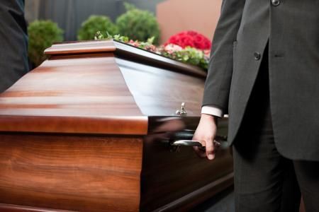 Wooden casket being carried by a person in a black suit at an outdoor funeral