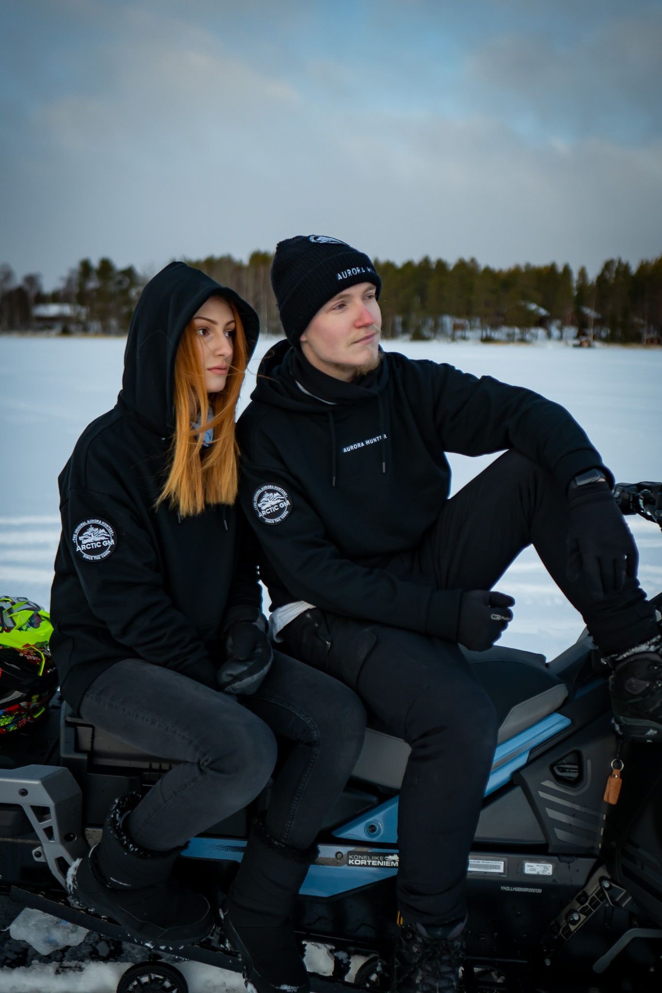 Man and woman wearing Arctic GM hoodies, sitting outdoors in snowy Lapland