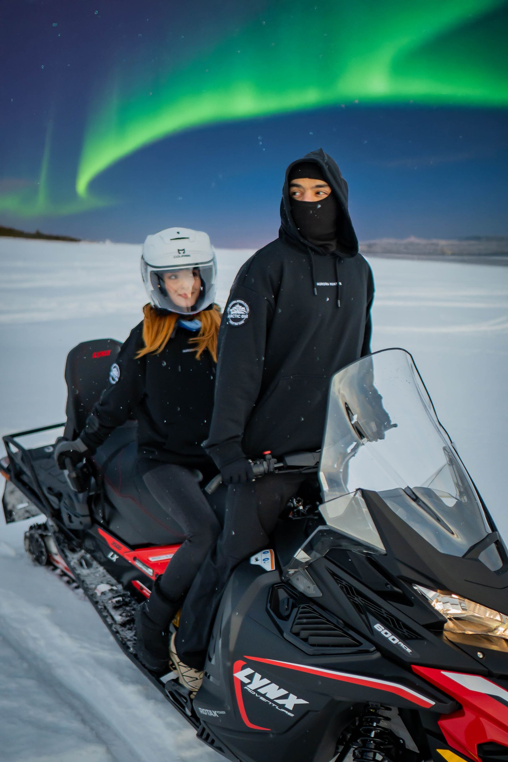 Couple riding snowmobile in Arctic GM outfits under Northern Lights in Finland
