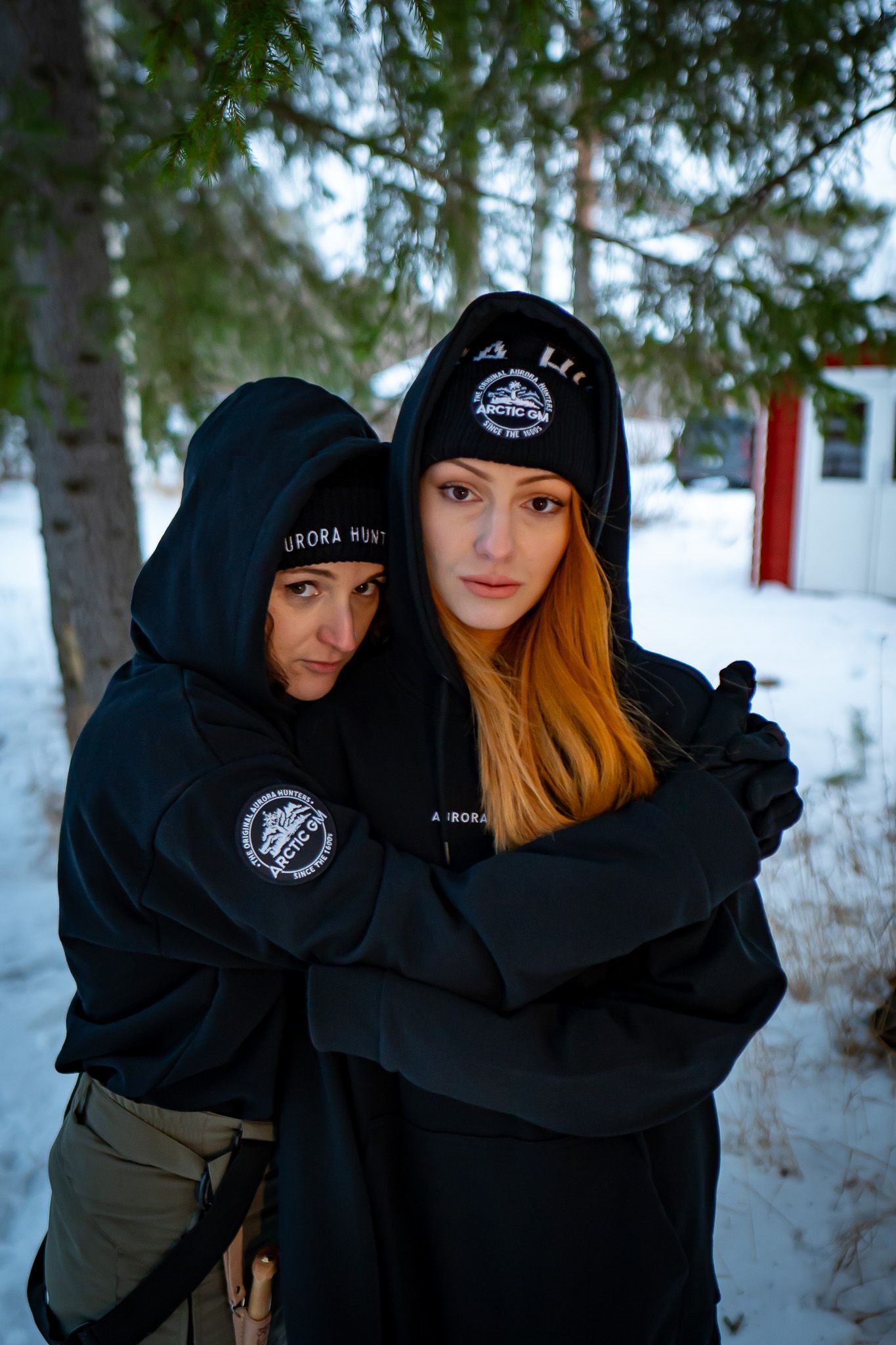 Two women hugging in matching Arctic GM hoodies and beanies, winter background