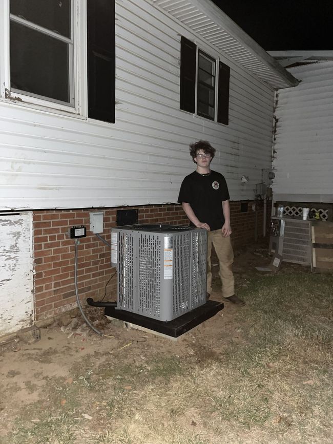 Person standing beside an outdoor HVAC unit next to a white house at night