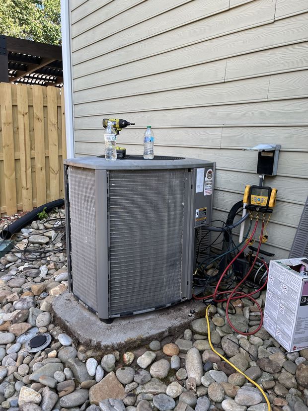 Outdoor air conditioner unit beside a house wall, with tools and cords nearby on rocky ground.