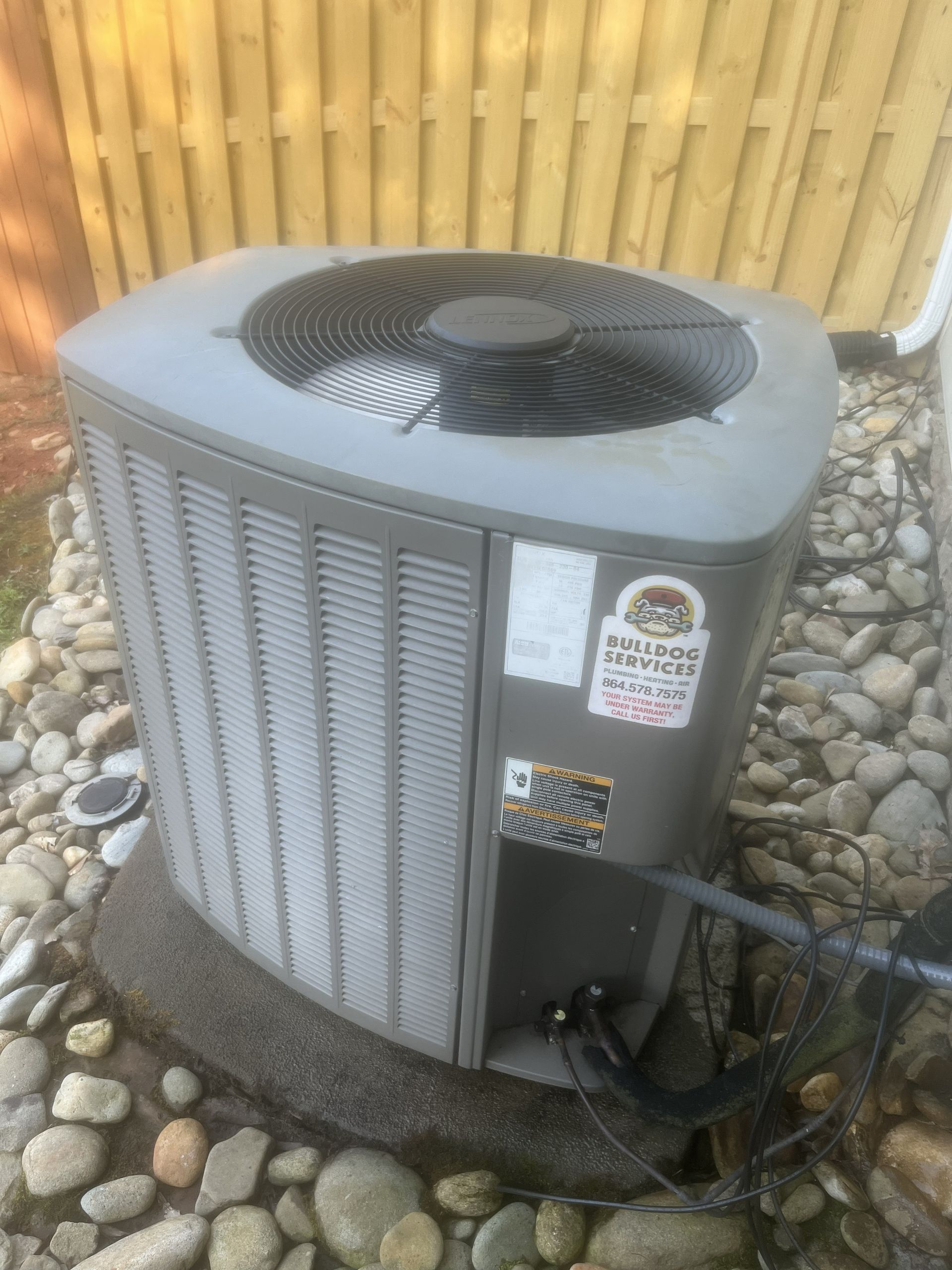 Outdoor central air conditioning unit beside a wooden fence and rocks
