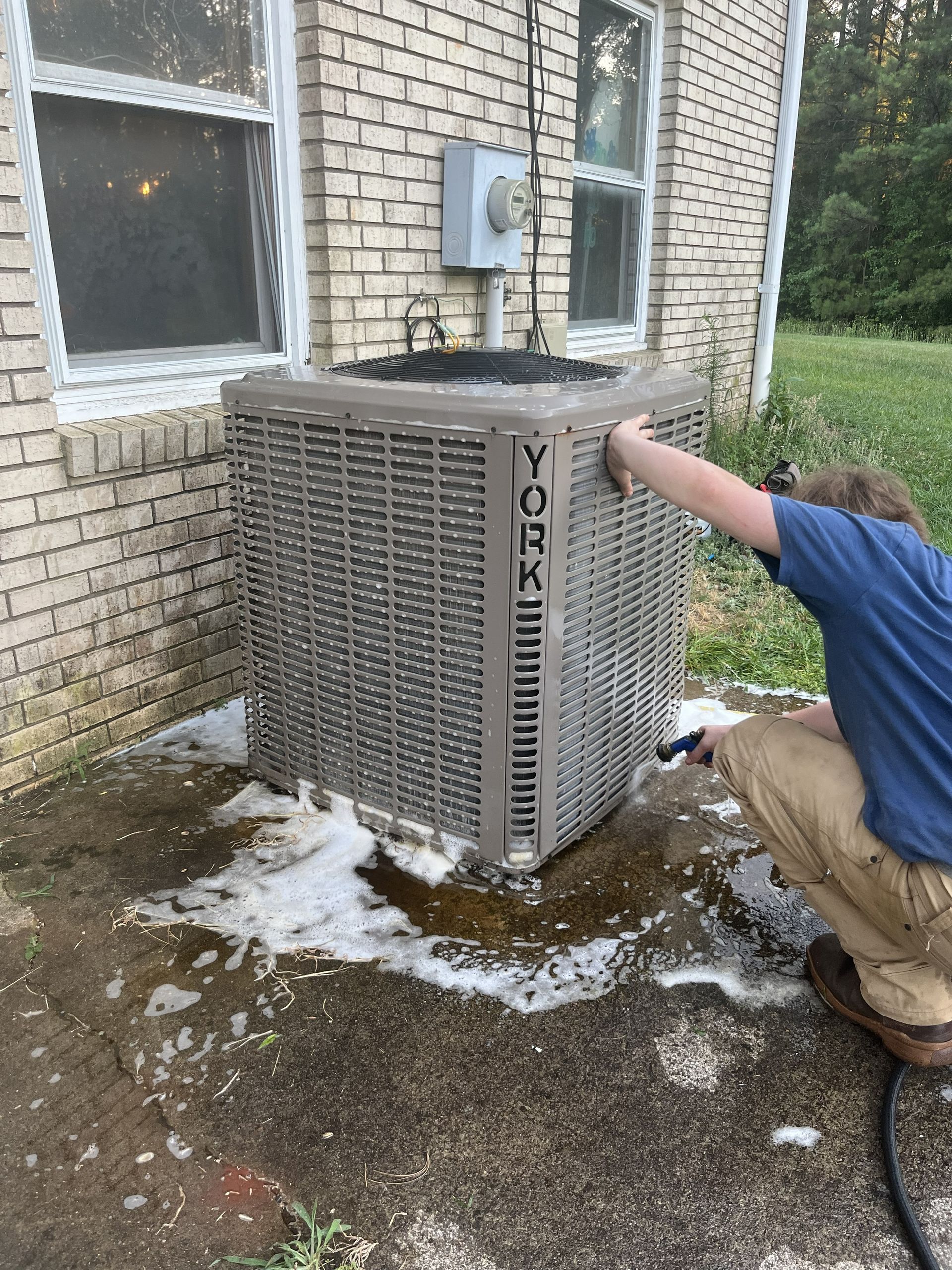 Technician kneels beside a YORK air conditioner unit outside a brick building, inspecting it.