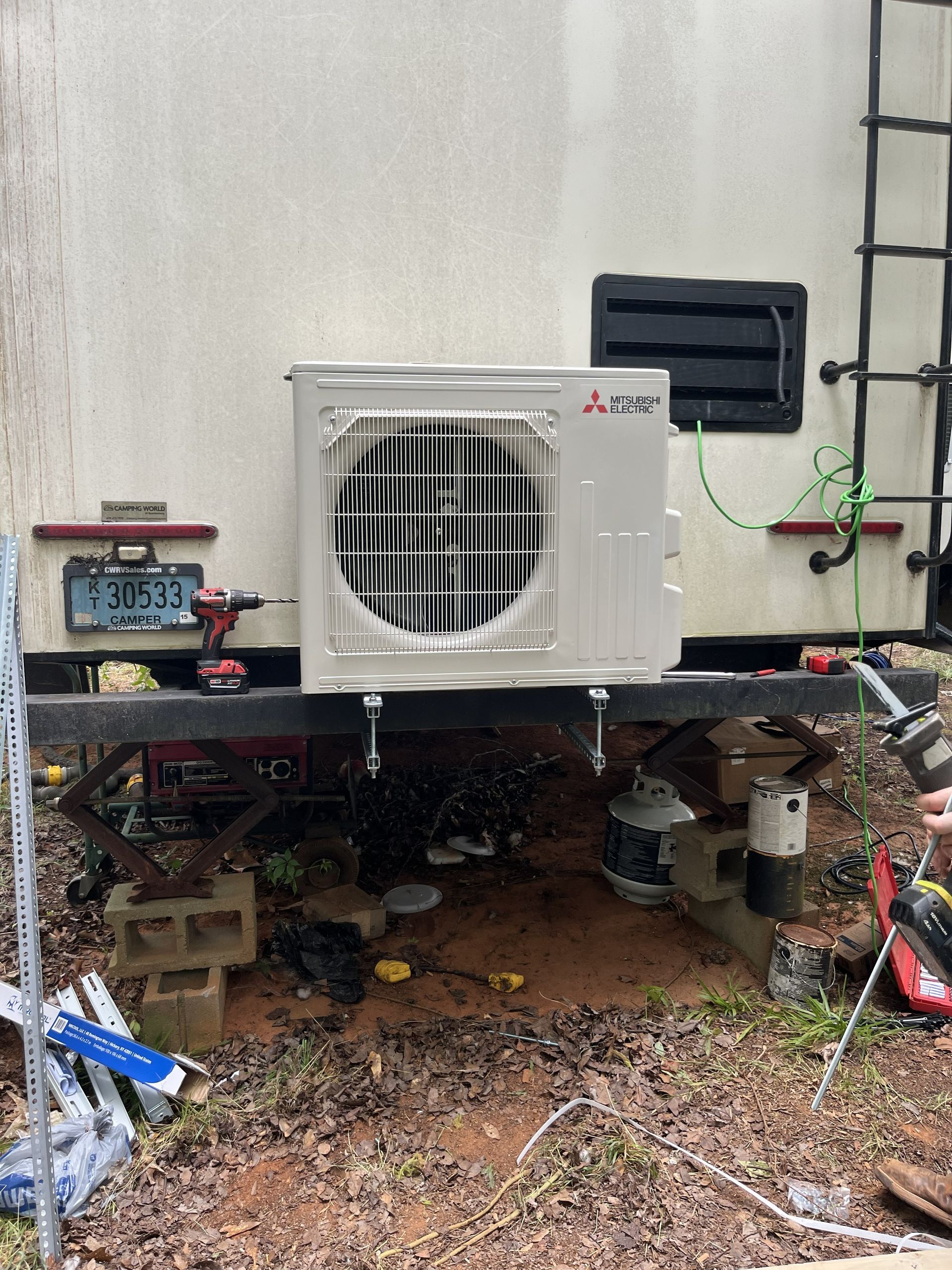 Outdoor HVAC unit mounted on a platform beside a building wall, with exposed pipes and a ladder nearby.