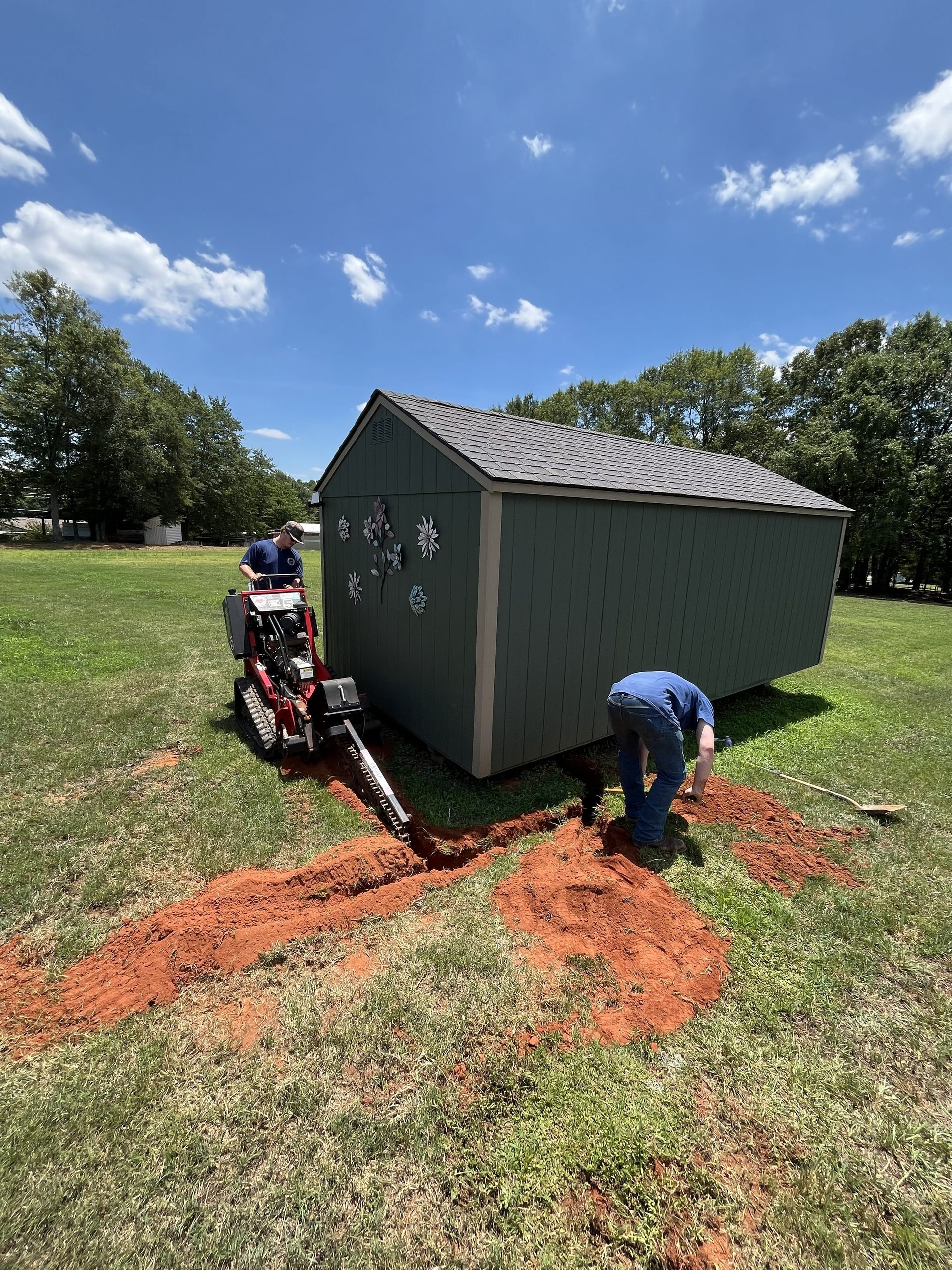 Two workers installing a small gray shed on red dirt in a grassy field under a blue sky.