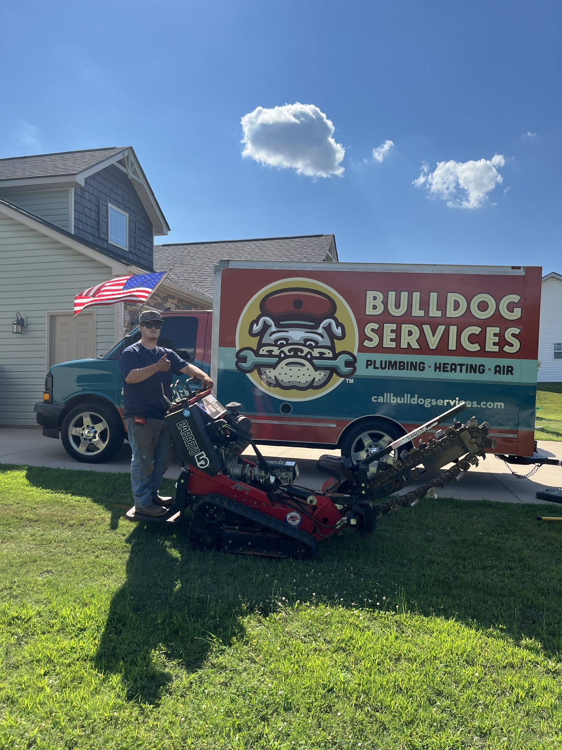 Man standing beside a red lawn mower and “BULLDOG SERVICES” trailer in a sunny suburban yard.