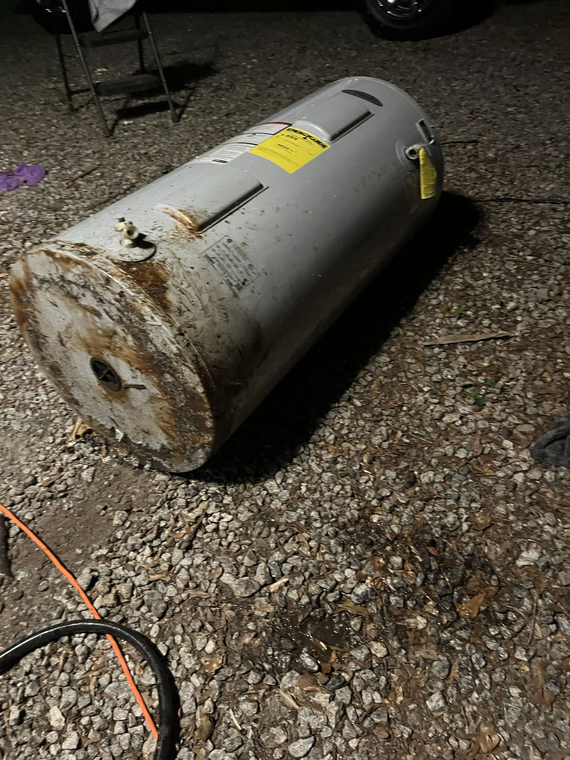 Rusty cylindrical metal tank lying on gravel at night, with yellow warning labels.