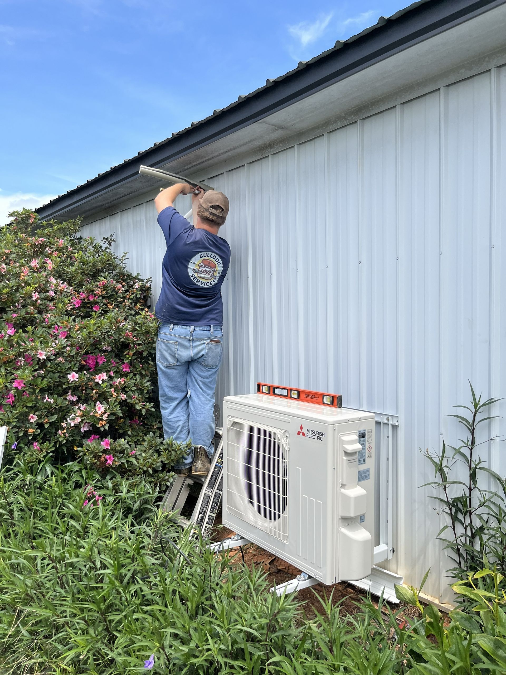 Worker cleaning gutters beside a house, standing on a ladder near an outdoor AC unit.