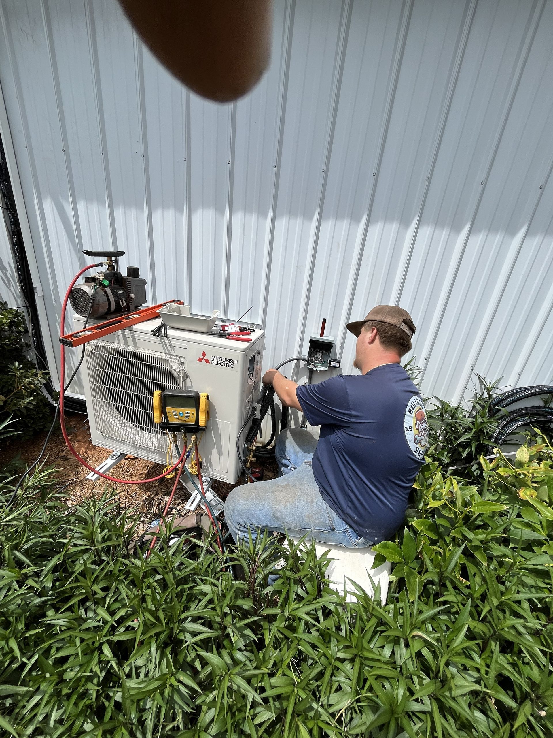 Man kneeling beside outdoor HVAC unit, working with tools against a corrugated metal wall.