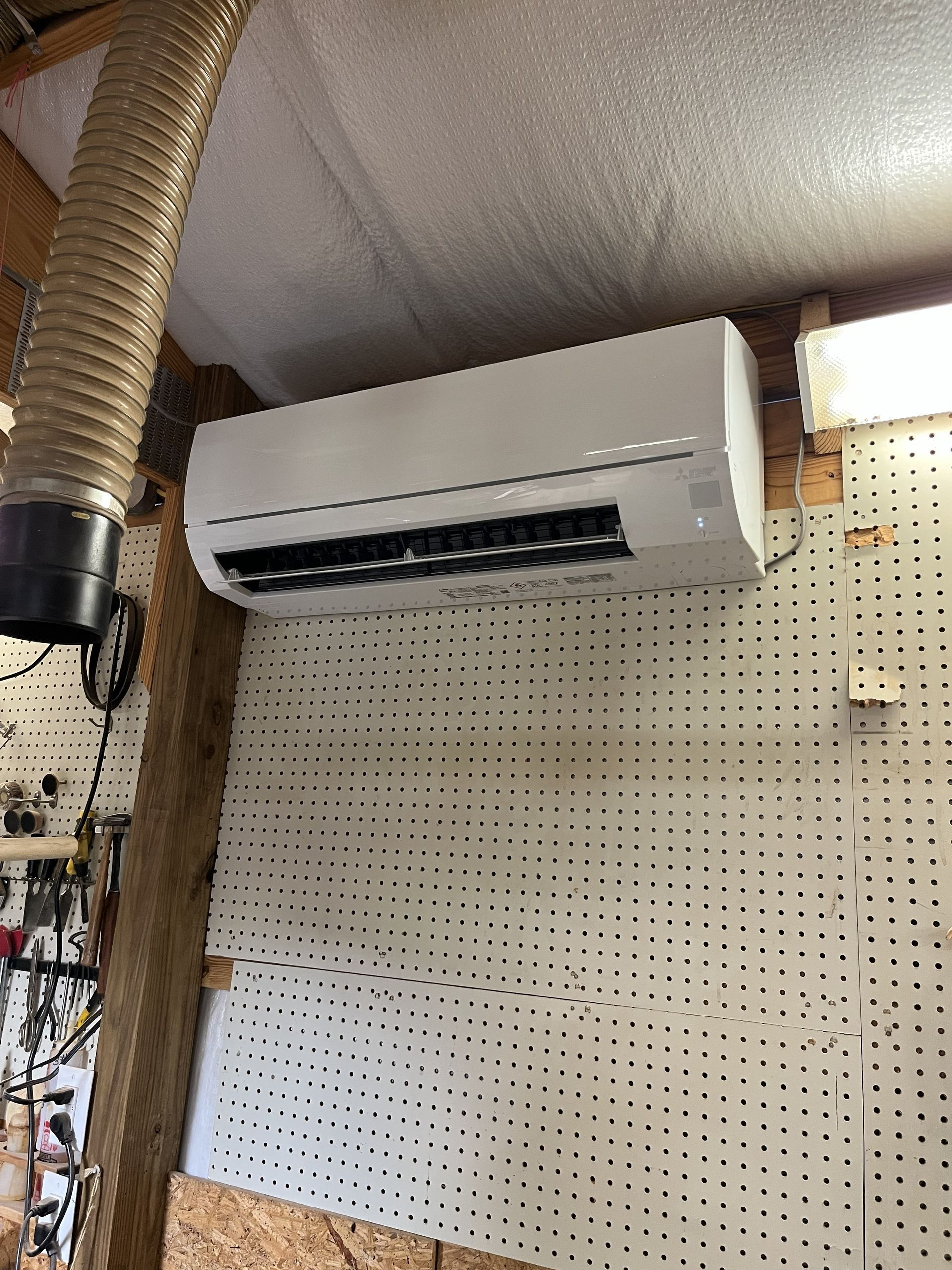 Wall-mounted white air conditioner above a pegboard wall with exposed ducting.