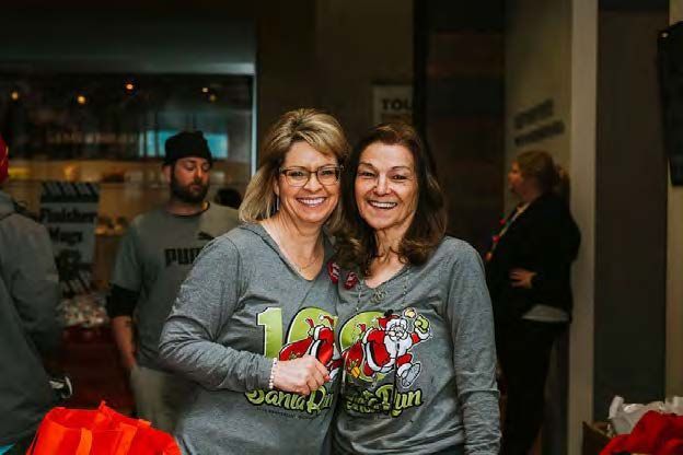 Two women smile, wearing matching Santa Run t-shirts. Others stand inside a building.